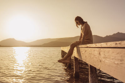 Man sitting on pier over lake against sky during sunset