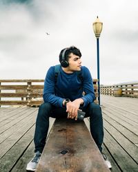 Full length of man listening music while sitting on bench against sky
