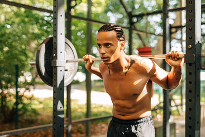 Shirtless man holding barbell while standing outdoors