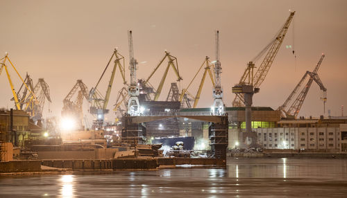 Cranes at harbor against sky during sunset