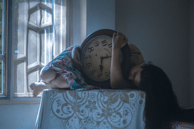 Young woman holding clock while lying on table at home
