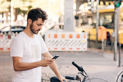 Young man using mobile phone while standing by bicycle on street in city