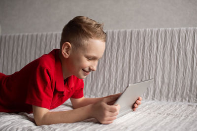 A cute boy in a red t-shirt is lying and playing on a tablet on the couch and laughing
