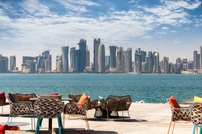 Chairs by river and buildings against sky in city