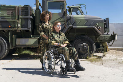 Young female military soldier with disabled colleague on wheelchair against truck at army base during sunny day