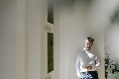 Businessman using digital tablet while leaning on wall at office