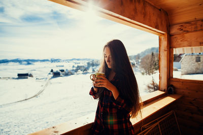 Portrait of young woman holding cup while standing by window in cottage during winter