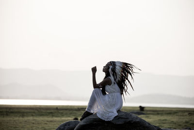 Side view of mid adult woman in traditional clothing sitting on rock against sky