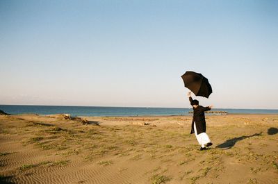 Full length of woman with umbrella standing on one leg at beach against clear sky