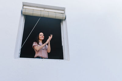 Young brunette woman with mask clapping from the window of the house with white background