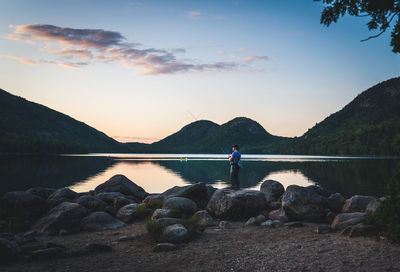 Man on rocks by lake against sky during sunset