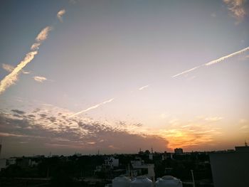 Vapor trails over silhouette city buildings against sky during sunset