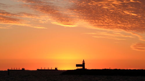 Silhouette lighthouse by sea against orange sky