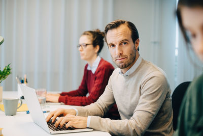 Portrait of confident businessman using laptop while sitting amidst female colleagues at creative office