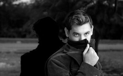 Portrait of man covering mouth with collar while standing by glass window