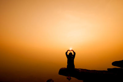 Silhouette person making heart shape on cliff against sky during sunset