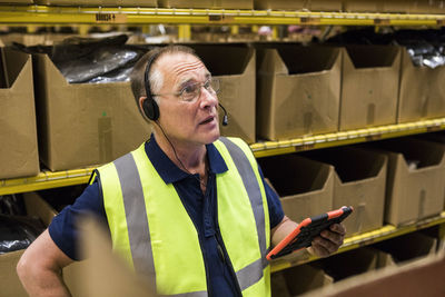 Senior male worker with digital tablet looking away while talking on headset against rack at distribution warehouse