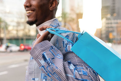 Crop anonymous smiling male customer with colorful shopping bags walking on paved walkway against tall residential buildings on street of city