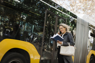 Smiling woman talking on smart phone and reading diary by bus