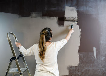 Woman climbed a ladder and catching a paint roller full of grey painting on a brown wall. painter is upping and downing roller covering the wall with grey painting what remains wet. horizonta