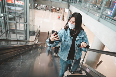Woman wearing flu mask standing on escalator holding smart phone at mall