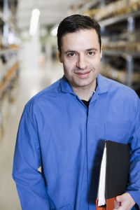 Front view portrait of confident male electrician holding file while standing in industry