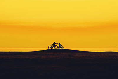 Silhouette friends riding bicycle on hill against orange sky