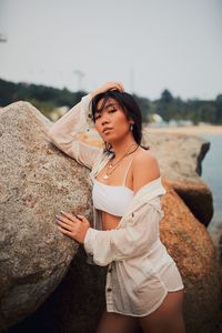 Portrait of young woman wearing bikini standing by rocks near sea against clear sky