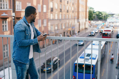 Young man standing with mobile phone on footbridge while looking at city street