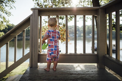 Rear view of baby boy standing by wooden railing against lake