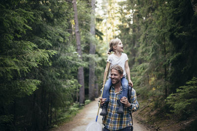 Smiling father carrying daughter on shoulder while walking in forest