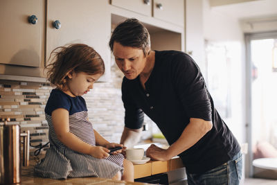 Daughter using phone while father standing in domestic kitchen