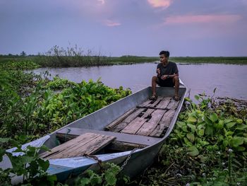 Man sitting on moored paddle boat in lake against sky during sunset