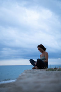 Side view of woman sitting on rock by sea against sky