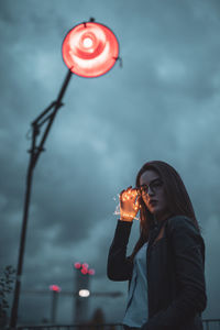 Low angle portrait of young woman holding string light while standing against cloudy sky at dusk