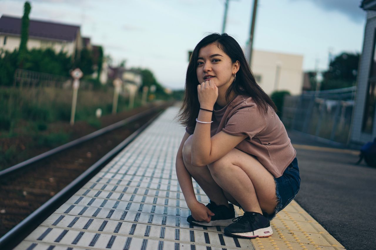 one person, real people, focus on foreground, lifestyles, young adult, track, transportation, leisure activity, full length, young women, rail transportation, railroad track, sitting, long hair, adult, beauty, hair, hairstyle, beautiful woman, outdoors