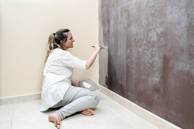 Blonde barefoot woman sitting on her floor starts painting a brown wall into a grey color with a small paintbrush. the woman wears a white shirt and a grey trousers. horizontal photo.