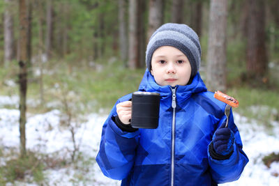 Portrait of cute boy having coffee and food while standing in forest during winter