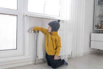 A small child in a sweater kneels and turns the thermostat on the heater in anticipation of heat