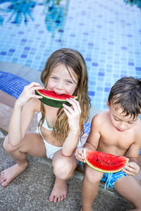 Siblings eating watermelon while sitting at swimming pool