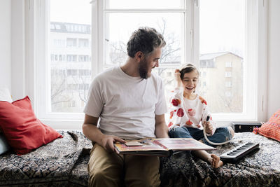 Happy man looking at daughter sitting with piano on bed at home