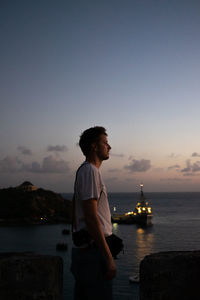 Side view of young man standing by sea against sky during sunset