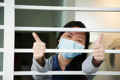 Portrait of young woman with face mask with thums up behind window