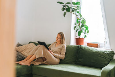 Beautiful blonde young woman artist illustrator drawing on tablet near window at home