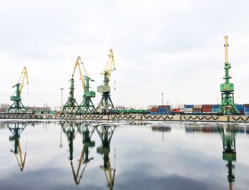 Reflection of cranes in lake at dock against cloudy sky
