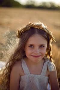 Girl child in a dress and a wreath on her head sit on a mown field of wheat at sunset in summer