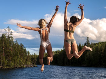 Full length of female friends jumping into lake against sky