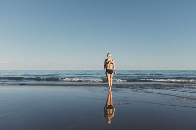 Rear view of woman wearing bikini while walking at beach towards sea against clear sky