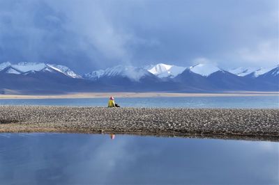 Rear view of person sitting on pebbles at lakeshore against snowcapped mountains