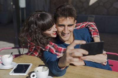 Smiling couple taking selfie while sitting on table at sidewalk cafe